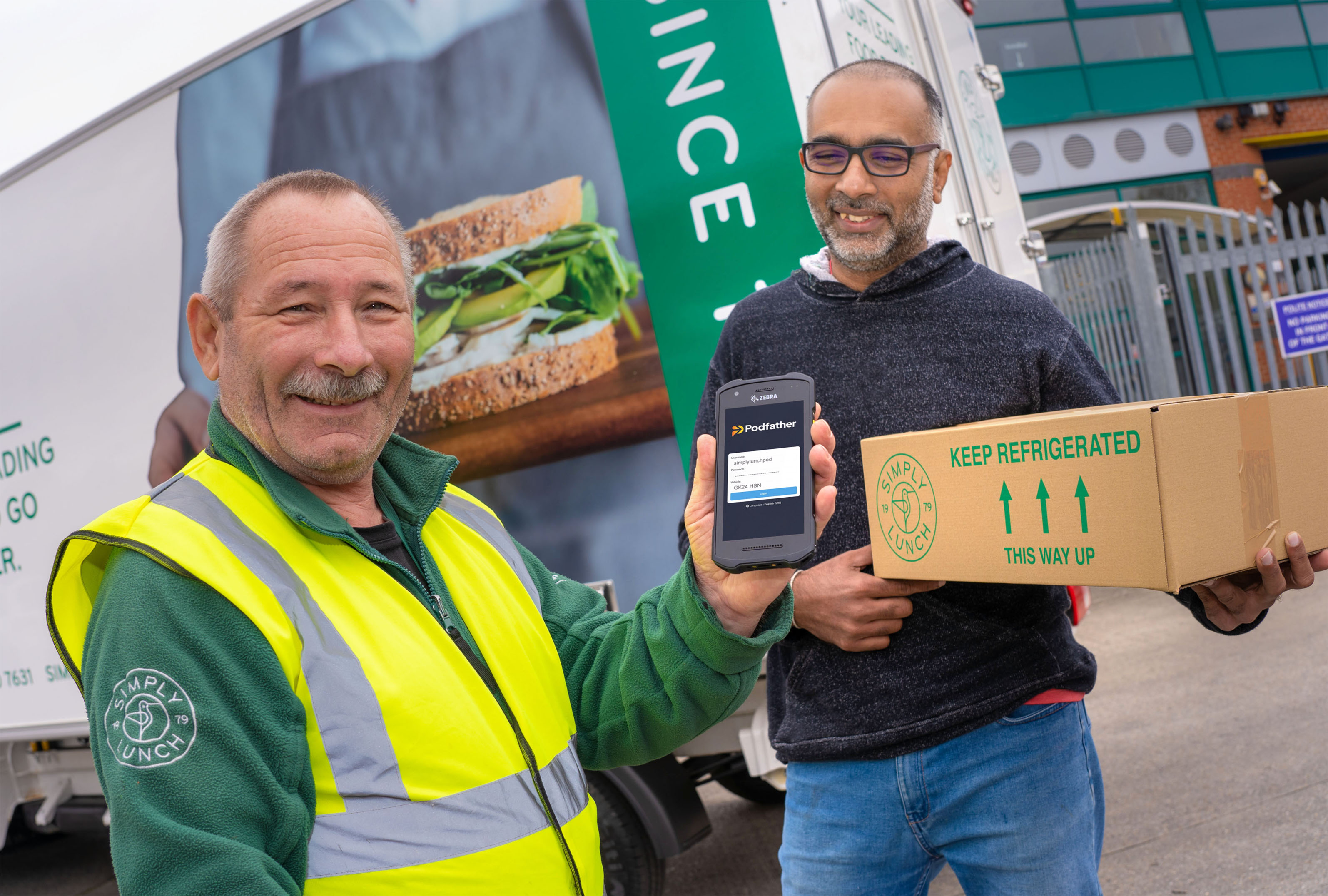 two men, one holding a box, one wearing a yellow high visibility vest, standing in front of a truck
