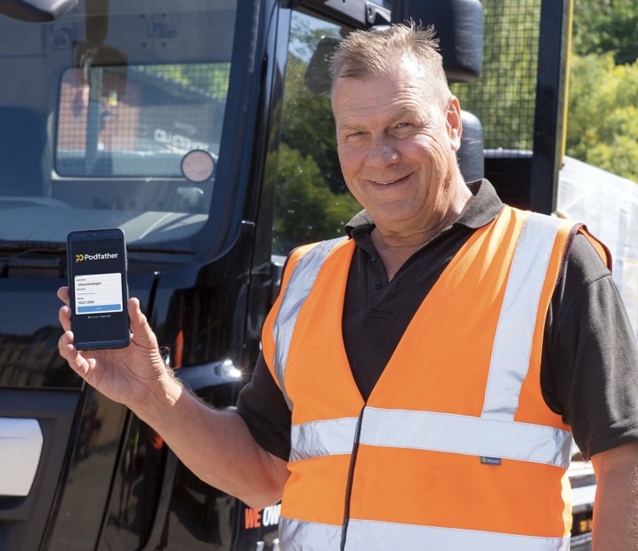Smiling man in an orange high vis vest standing in front of a truck holding a mobile phone